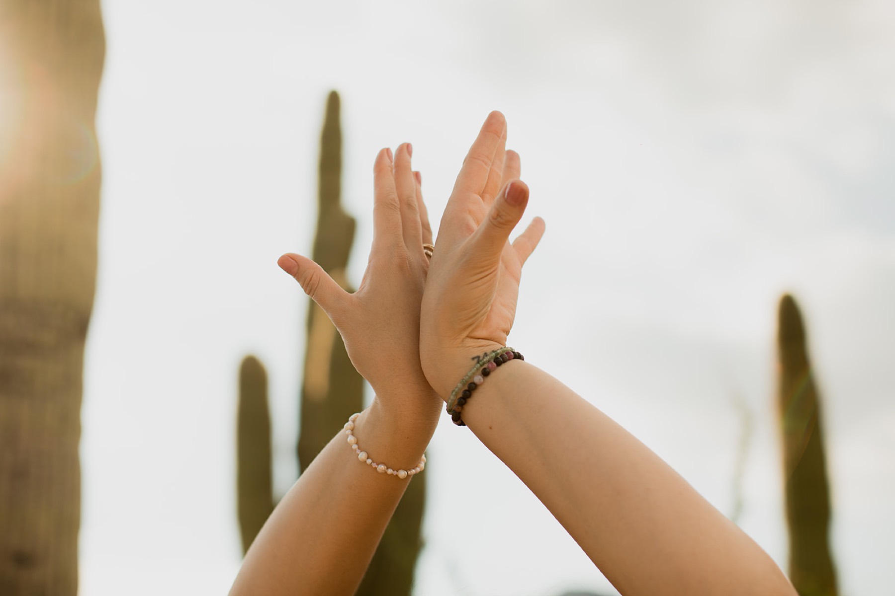 Two arms raised and crossed toward a bright sky between desert cacti, layered with beaded bracelets